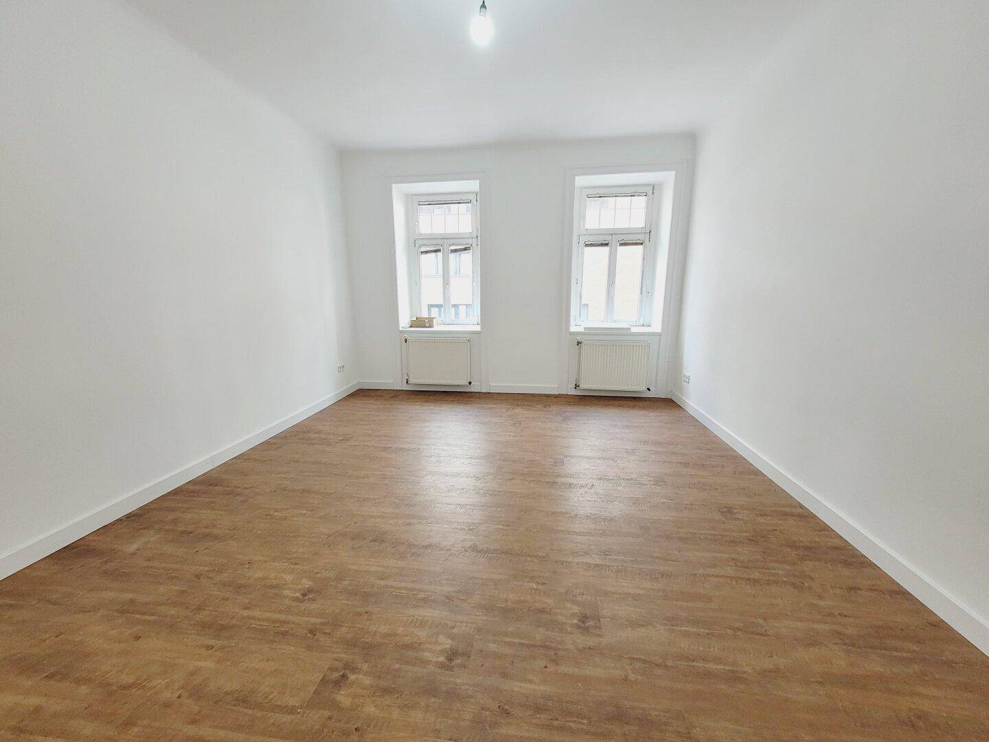 Lichtdurchflutetes Zimmer mit zwei großen Fenstern und neuem Holzboden, bietet viel Tageslicht.