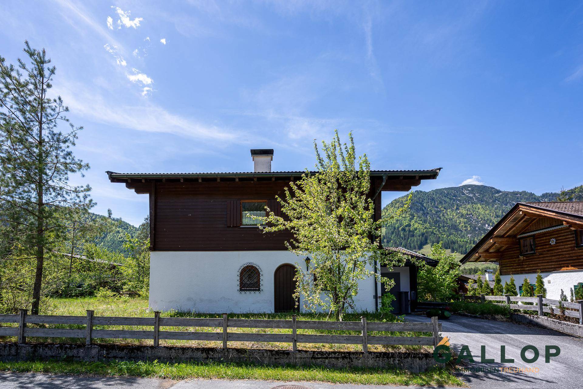 Seitenansicht des Hauses im alpinen Stil mit Holzzaun und einem jungen Baum im Vorgarten.