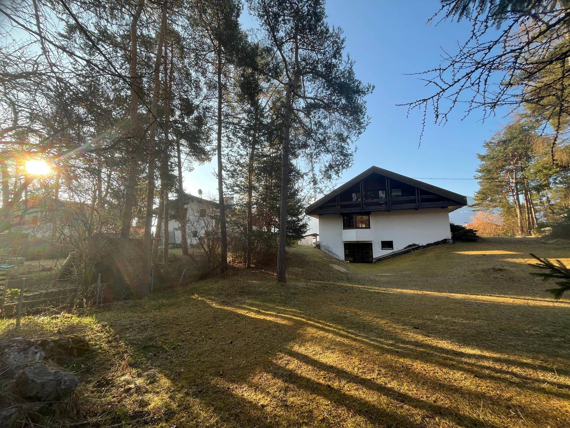 Hausfassade mit großem Rasenbereich und hohen Bäumen im Sonnenlicht.