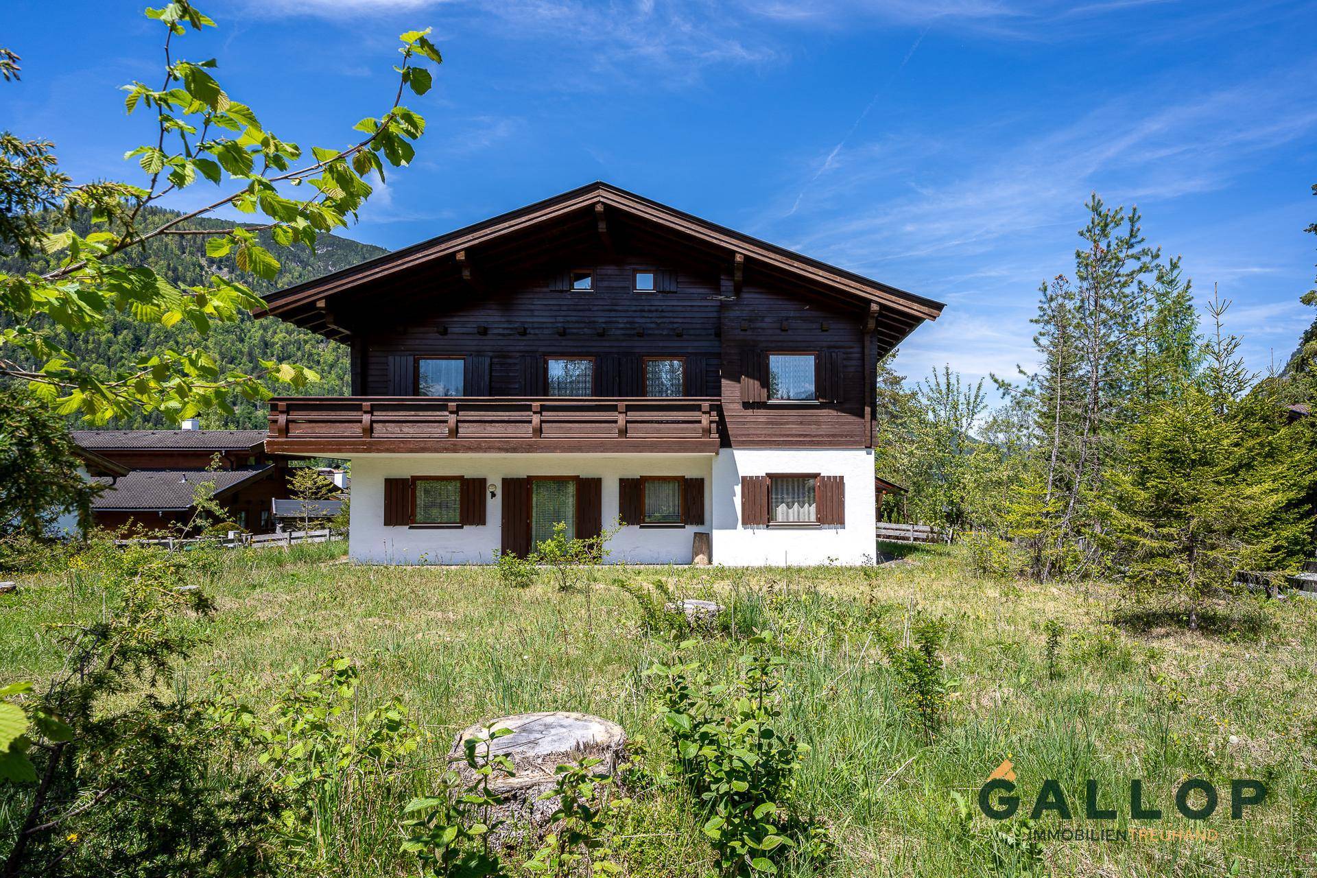 Vorderansicht des alpinen Hauses mit Holzbalkon und weißer Fassade, umgeben von Grünflächen.