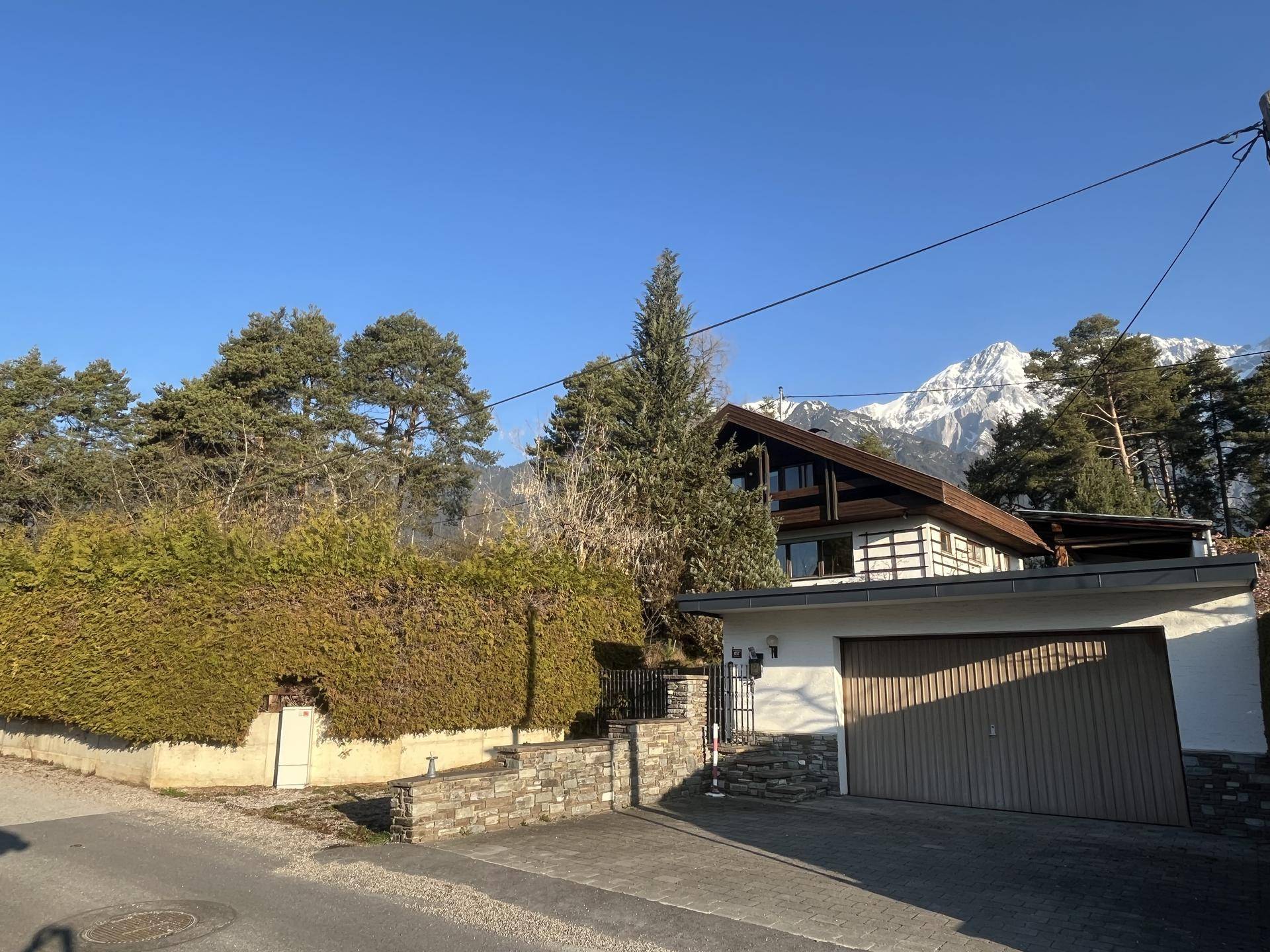 Vorderansicht des Hauses mit Garage, umgeben von einer Hecke und einem gepflasterten Bereich, mit Bergen im Hintergrund.
