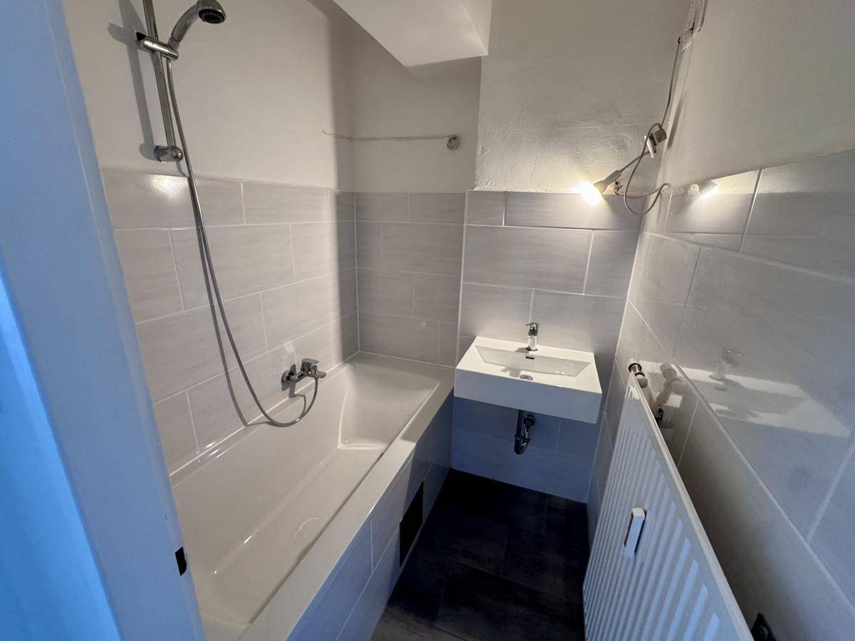Kompaktes Badezimmer mit Badewanne, Duschkopf, Waschbecken und modernen grauen Fliesen.