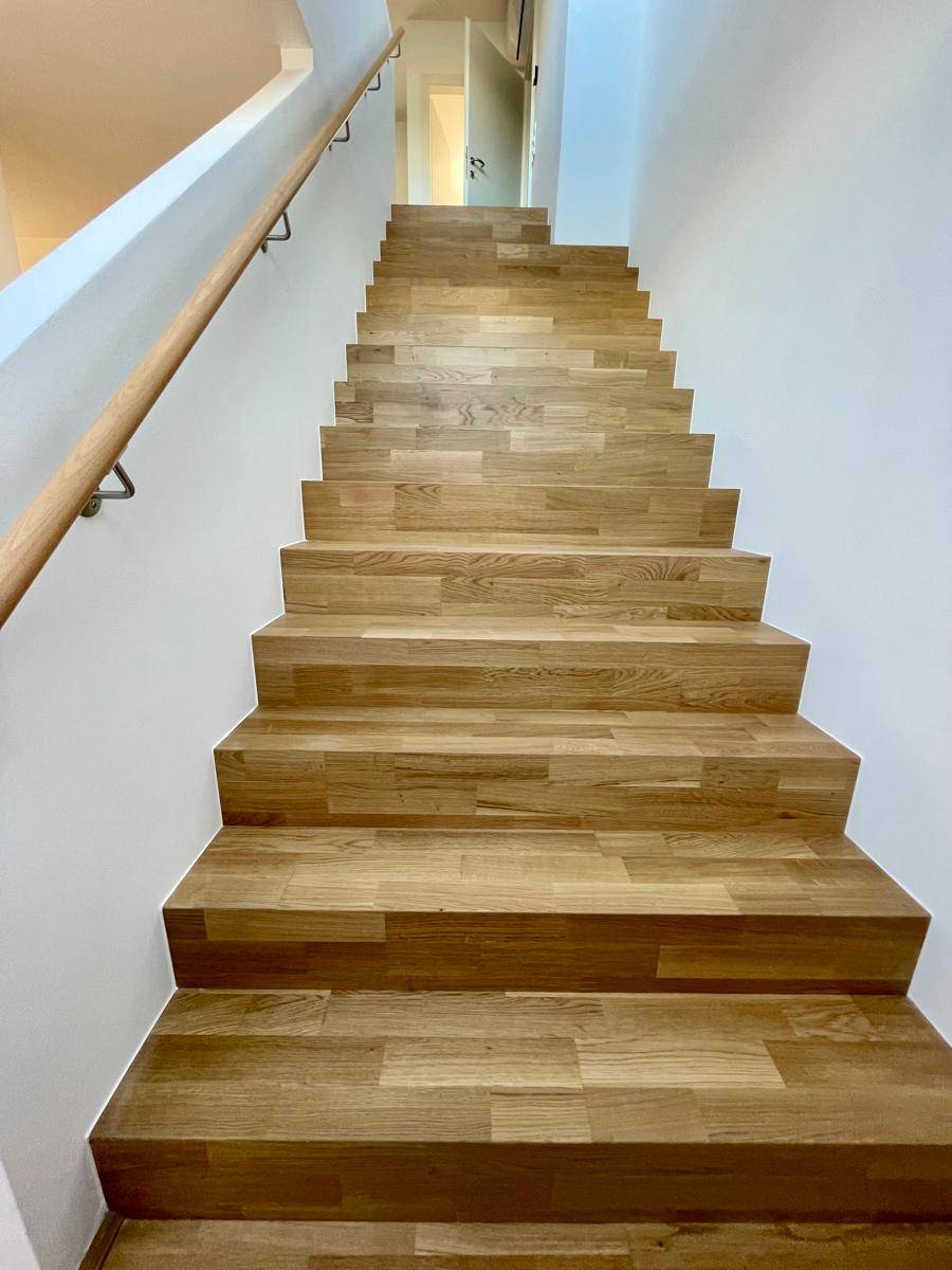 Moderne Holztreppe mit hellem Holzgeländer und weißen Wänden, die in die obere Etage führt.