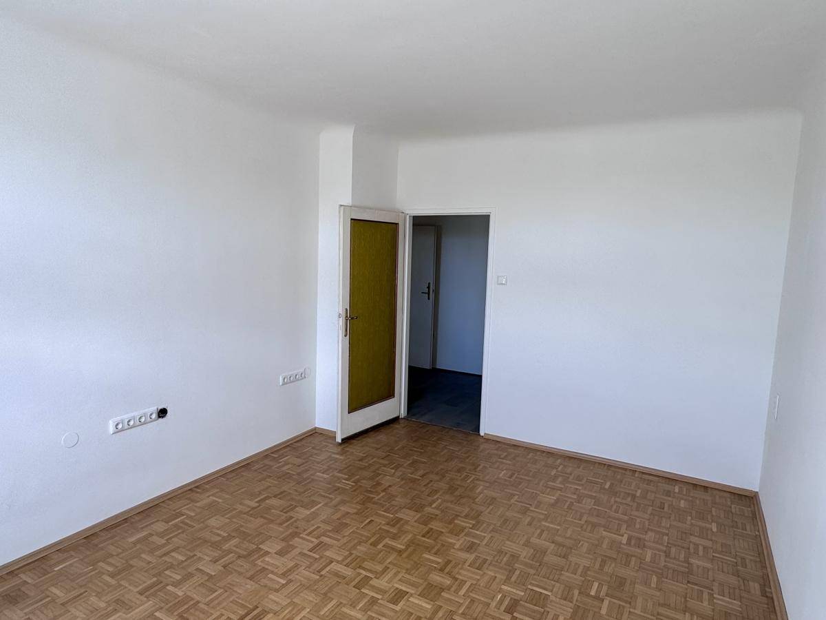 Geräumiges Zimmer mit Parkettboden und einer Tür mit gelbem Glaseinsatz.