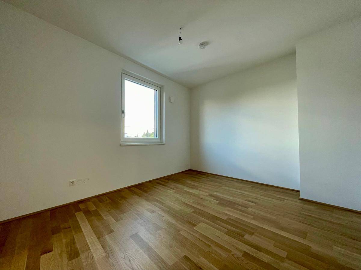 Einfaches, helles Zimmer mit Holzparkettboden und einem Fenster, ideal als Schlafzimmer.
