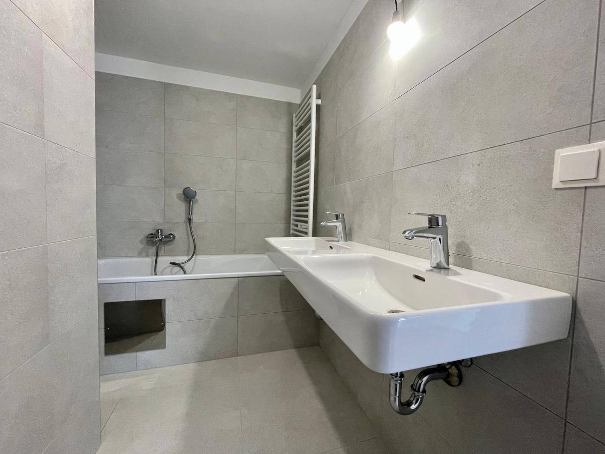 Modernes Badezimmer mit Doppelwaschbecken, Badewanne und hellgrauen Fliesen an Wänden und Boden.