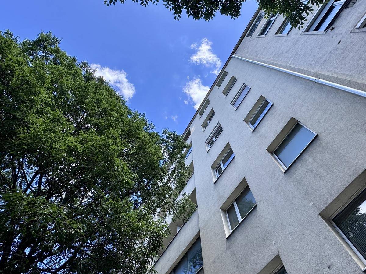 Fassade eines Gebäudes mit vielen Fenstern und grünen Bäumen unter blauem Himmel.