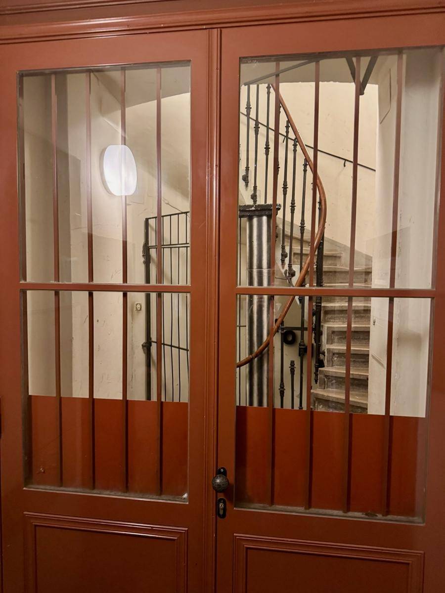 Blick durch verglaste Holztüren auf ein historisches Treppenhaus mit schmiedeeisernem Geländer.