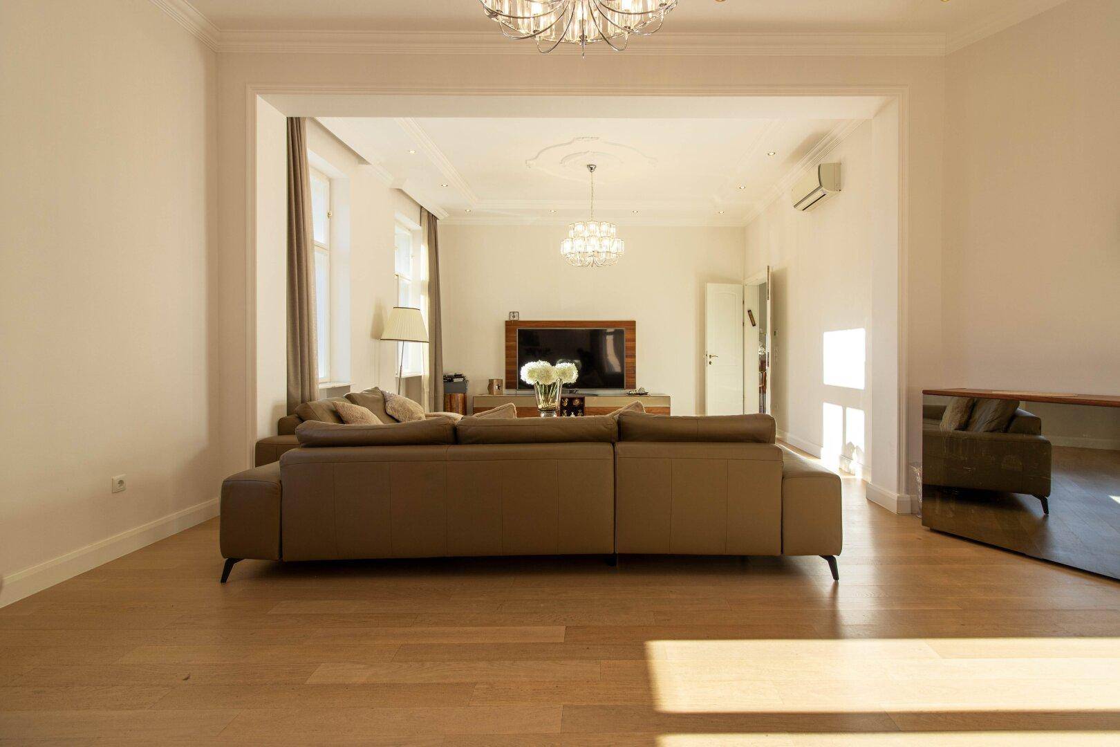 Geräumiges Wohnzimmer mit beigem Sofa, Parkettboden und Blick auf den Fernseher.