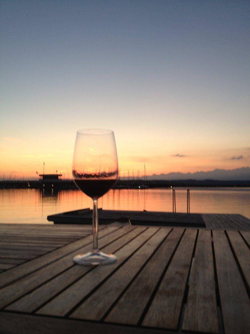 Romantischer Sonnenuntergang über dem See, eingefangen mit einem Weinglas auf einem Holzsteg.