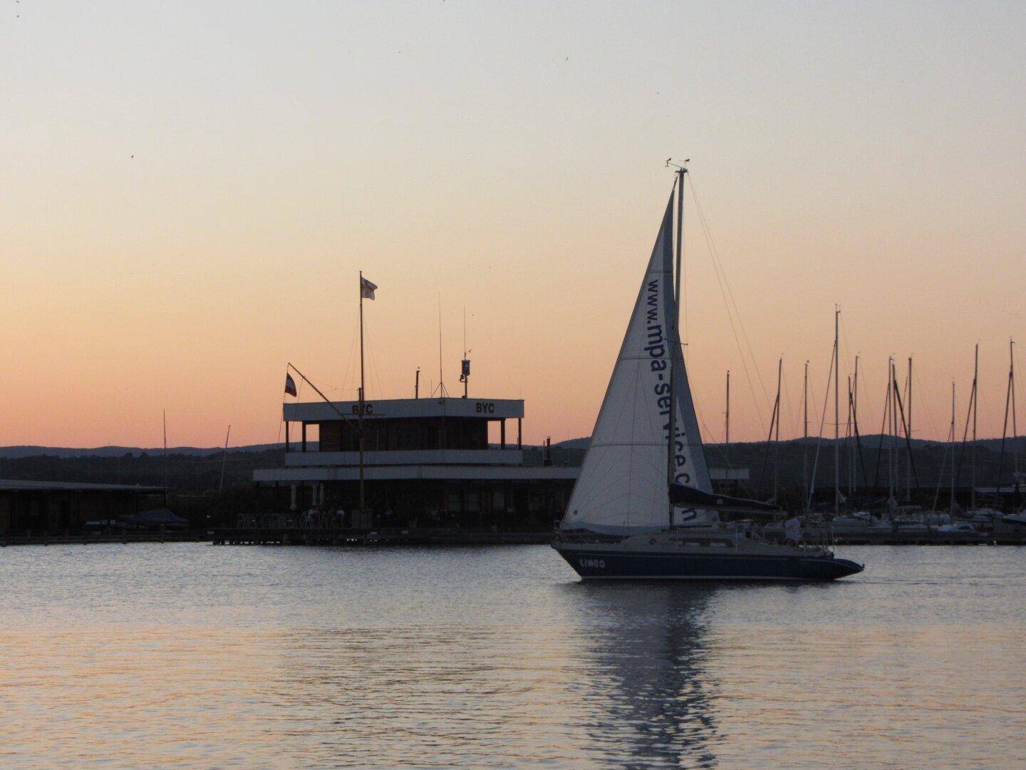 Segelboot auf dem See bei Sonnenuntergang mit einem Gebäude im Hintergrund.