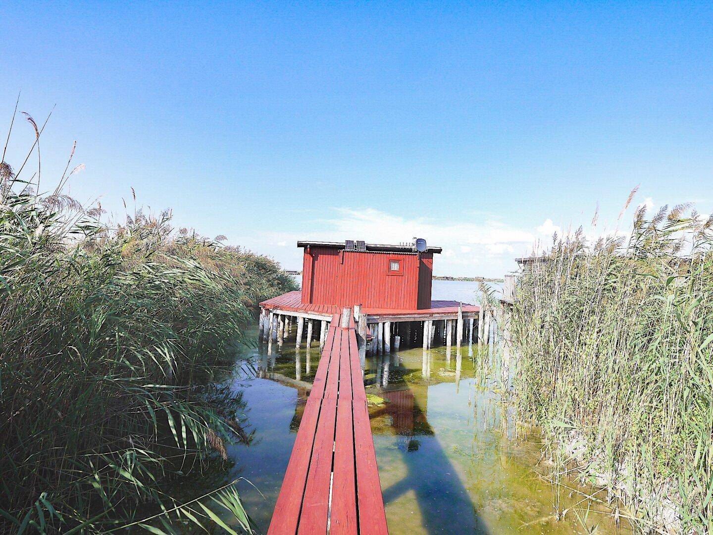 Rotes Holzhaus auf Stelzen im Wasser, erreichbar über einen langen Holzsteg durch das Schilf.