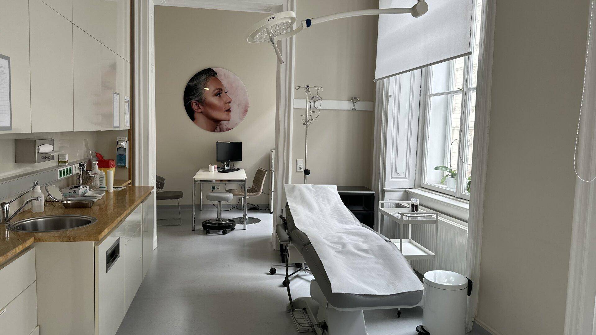 Behandlungsraum mit Liege, Schreibtisch, medizinischen Geräten und großen Fenstern.