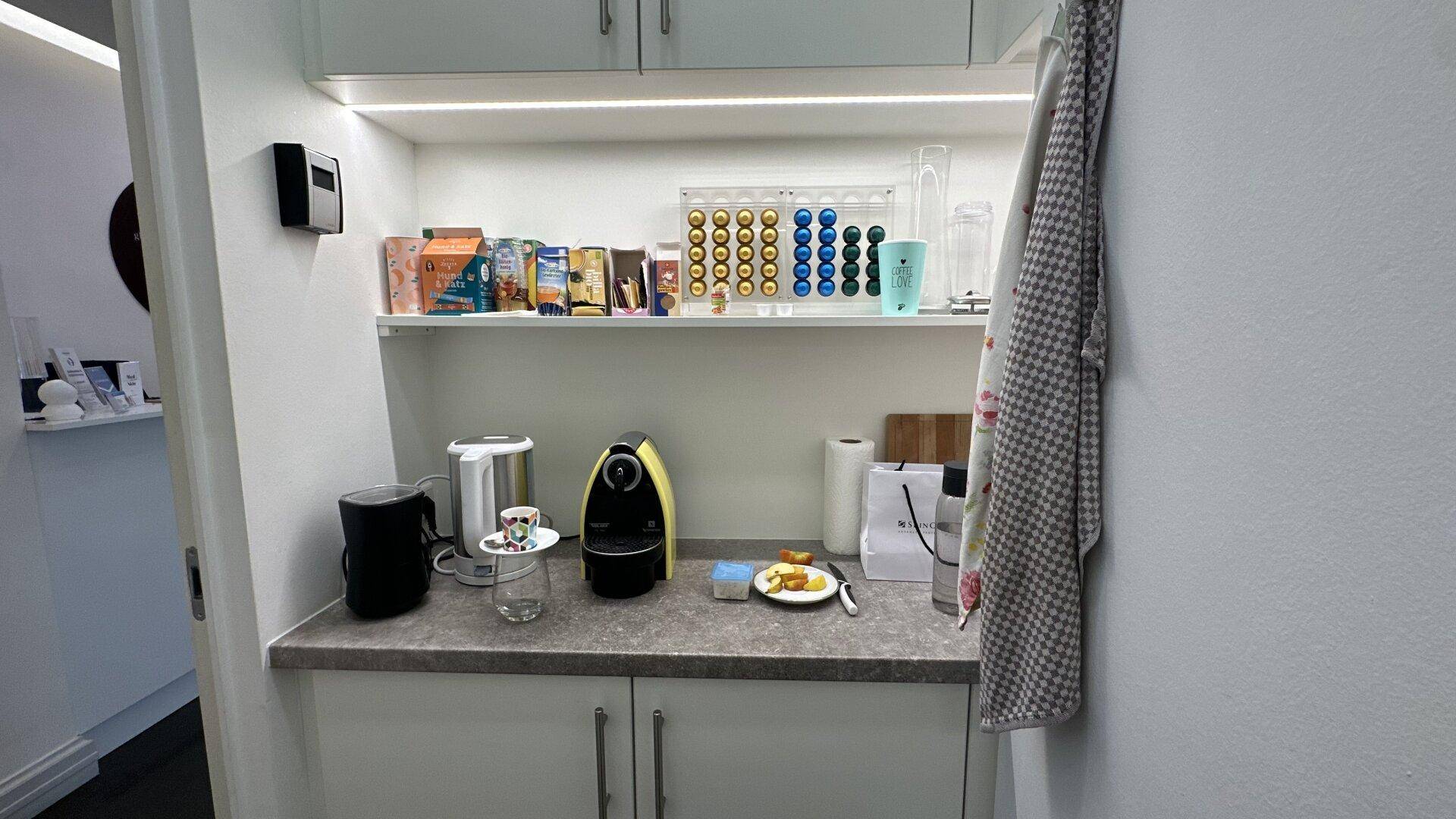 Kleine Küchenzeile mit Kaffeemaschine, Kapseln und Geschirr auf der Arbeitsplatte.