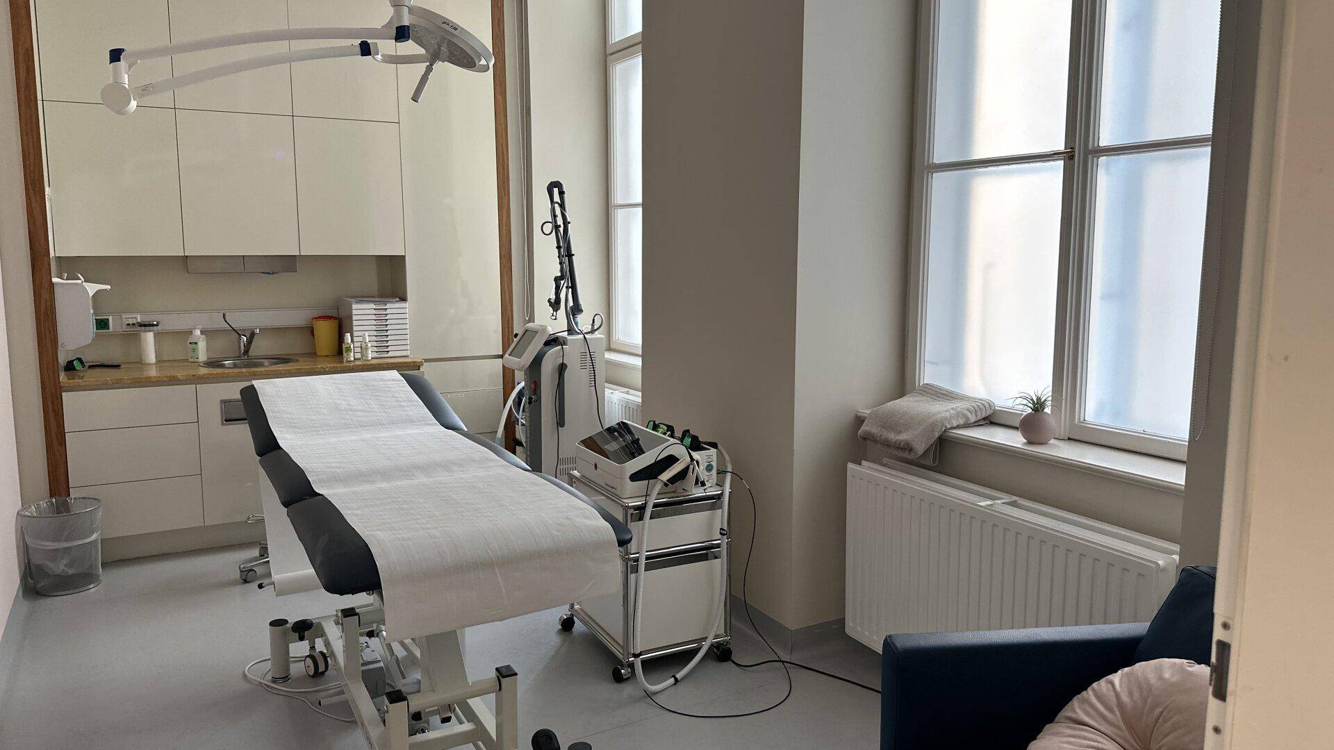 Behandlungsraum mit Liege, medizinischen Geräten und großen Fenstern.