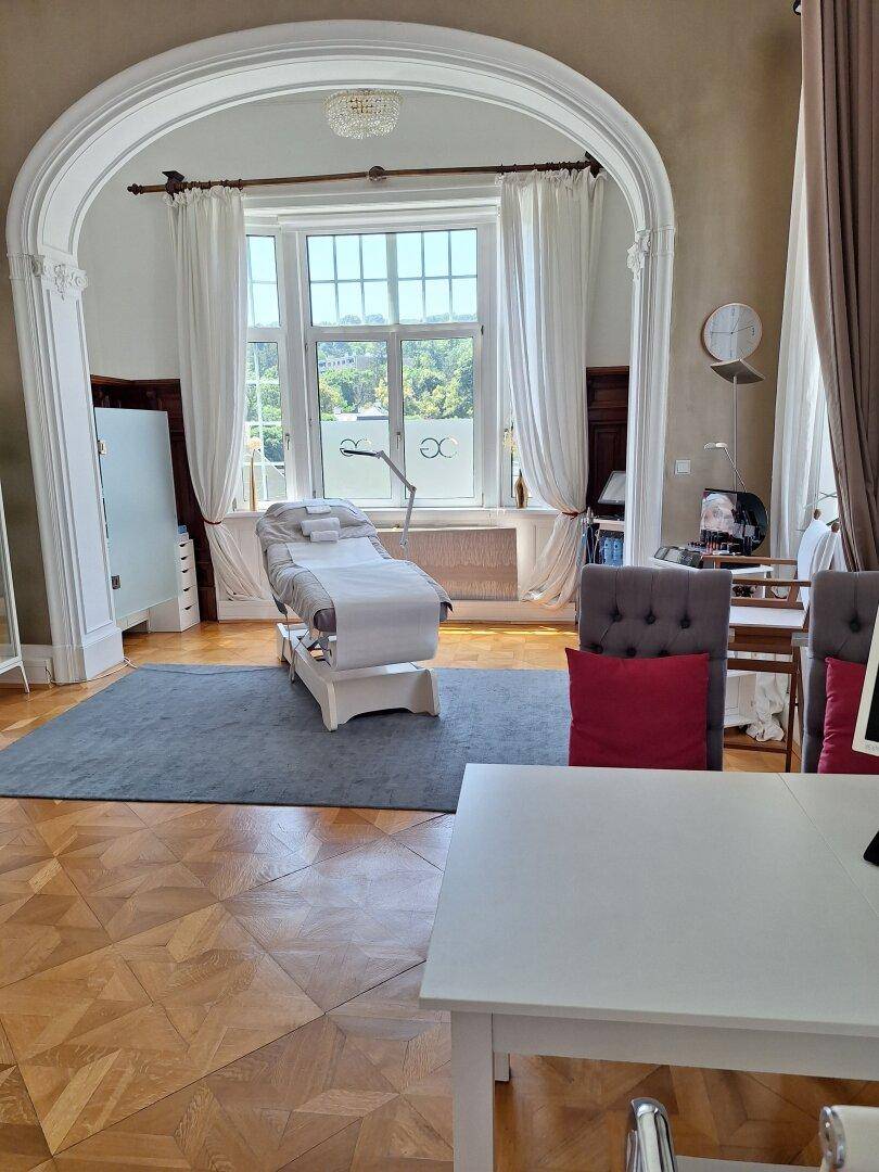 Eleganter Behandlungsraum mit Parkettboden, großen Bogenfenstern und einer medizinischen Liege.