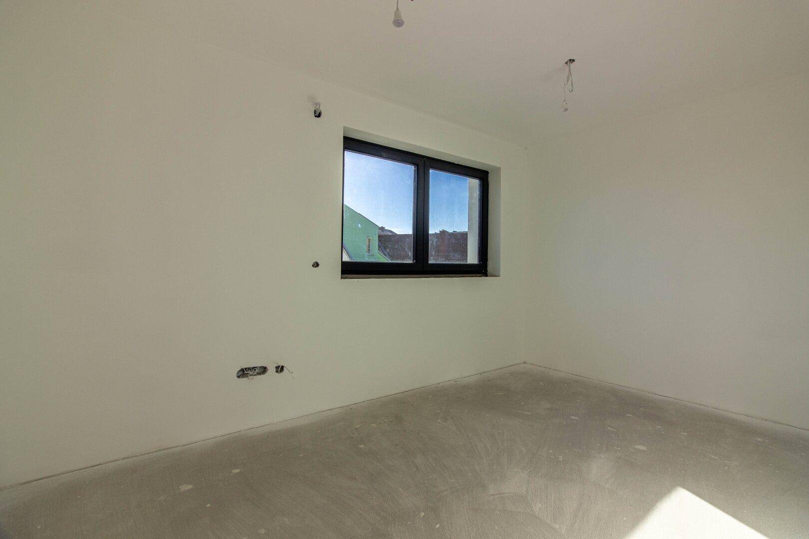 Heller Raum mit Betonboden und Fenster, das viel Tageslicht in das zukünftige Schlafzimmer bringt.