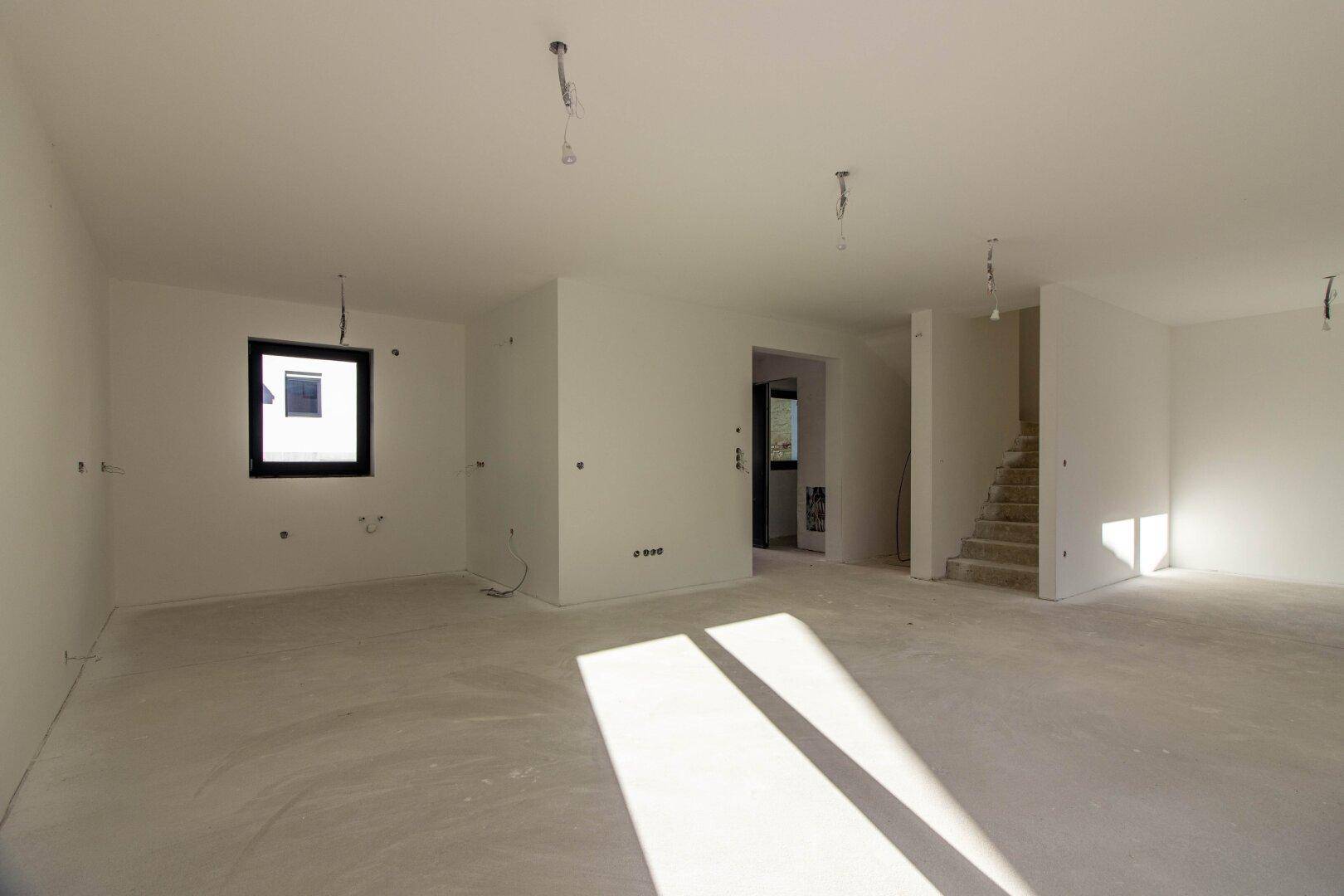 Großer, heller Raum im Rohbau mit Betonboden und Treppenaufgang, ideal für individuelle Gestaltung.