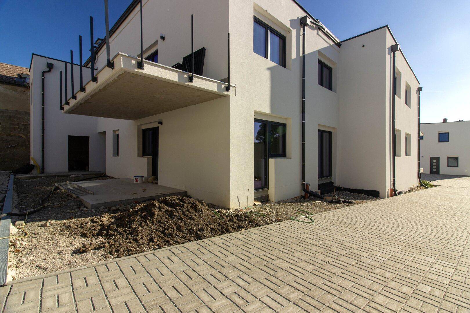 Seitenansicht eines Neubaus mit einem im Bau befindlichen Balkon und gepflastertem Zugangsbereich.