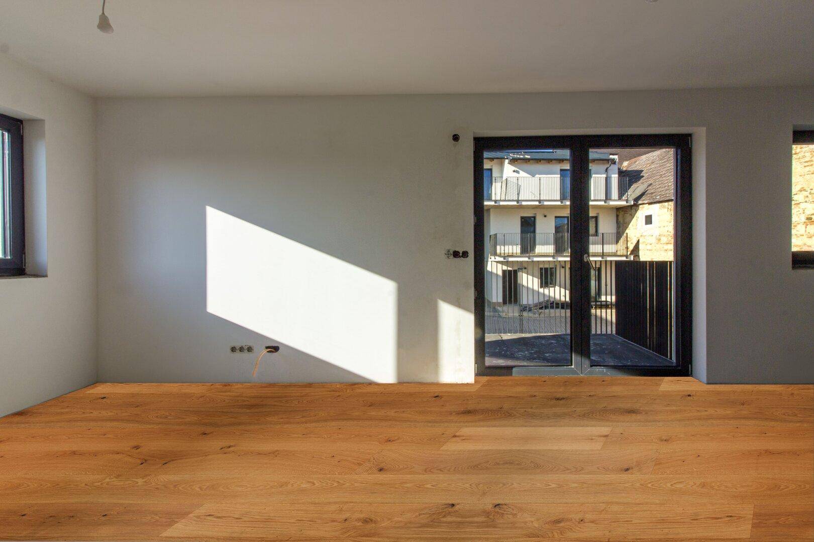 Lichtdurchfluteter Raum mit Holzboden und großer Glastür, die zu einem Außenbereich führt.