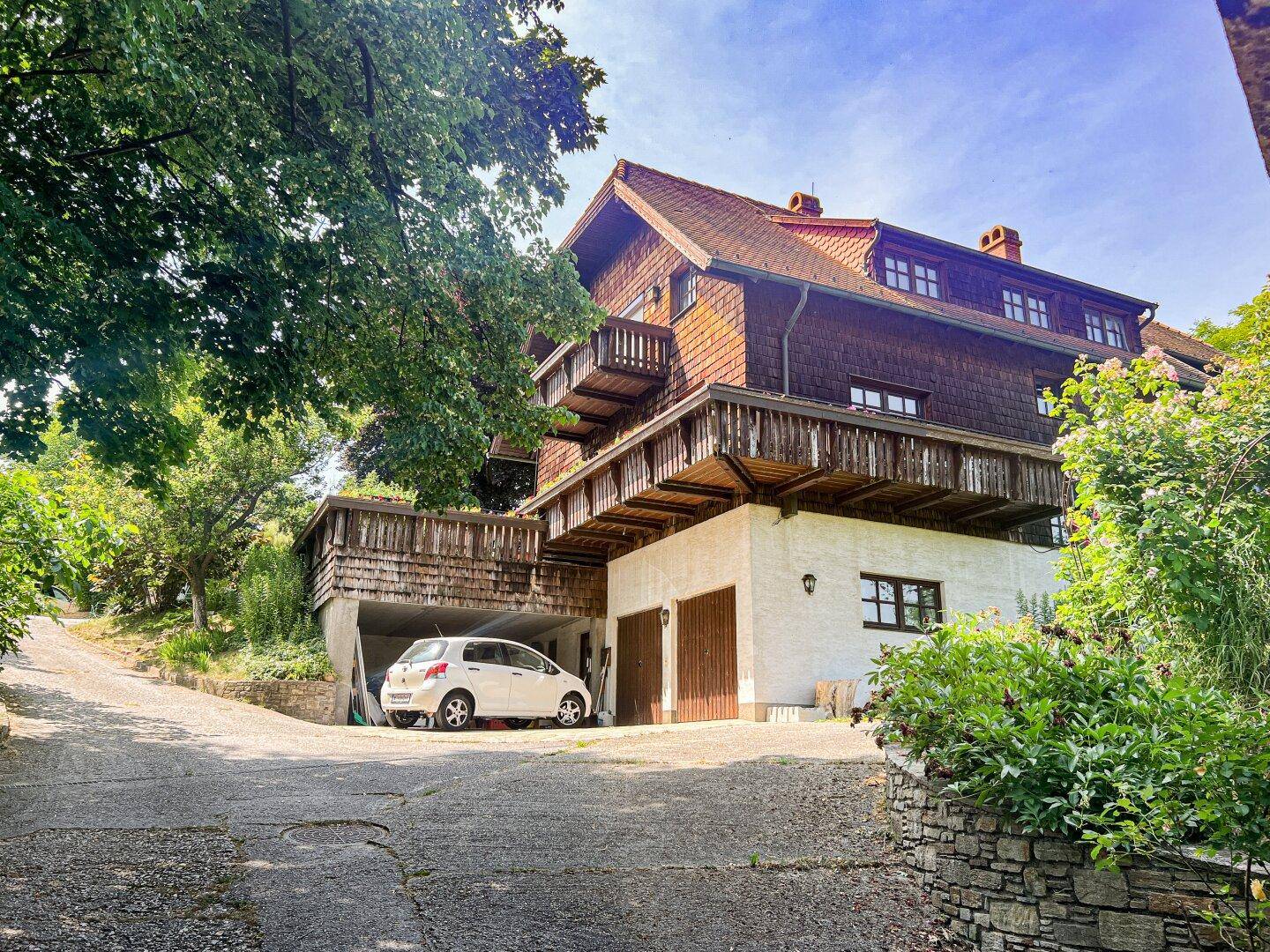Traditionelles Holzhaus mit integrierter Garage und Balkonen, umgeben von Bäumen und Grünflächen.