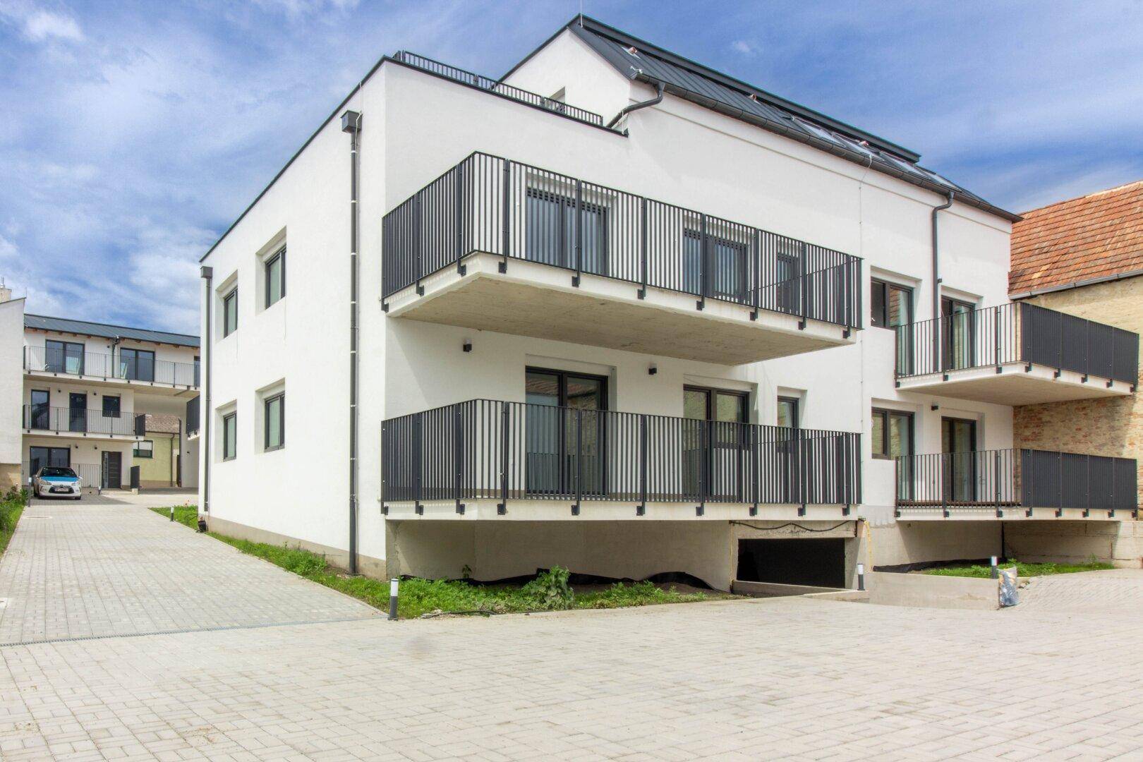 Ansicht eines modernen Wohnkomplexes mit mehreren Balkonen, weißer Fassade und Zufahrt zur Tiefgarage.