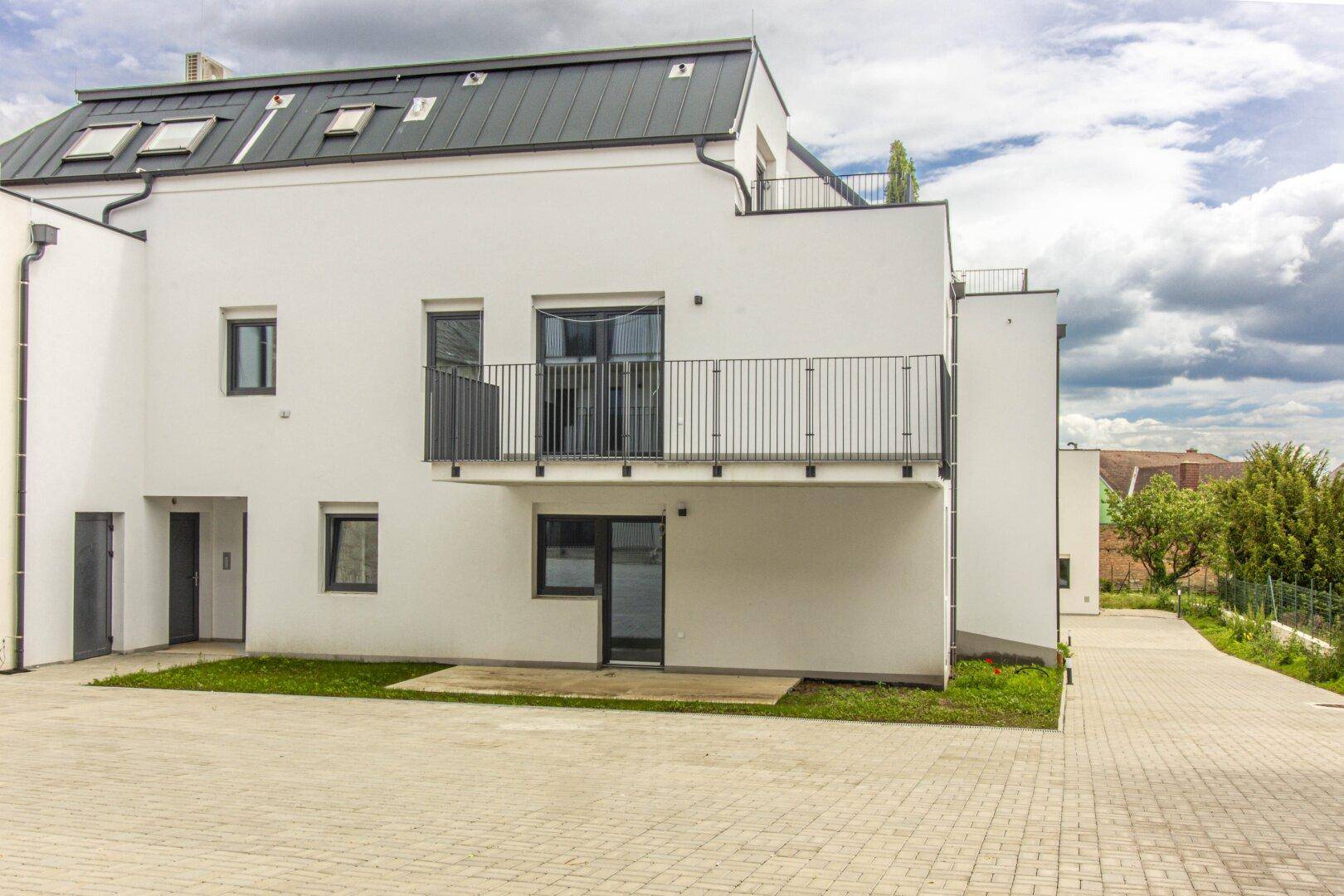 Moderne weiße Fassade eines Wohngebäudes mit dunklem Dach und Balkonen, umgeben von gepflasterten Flächen.