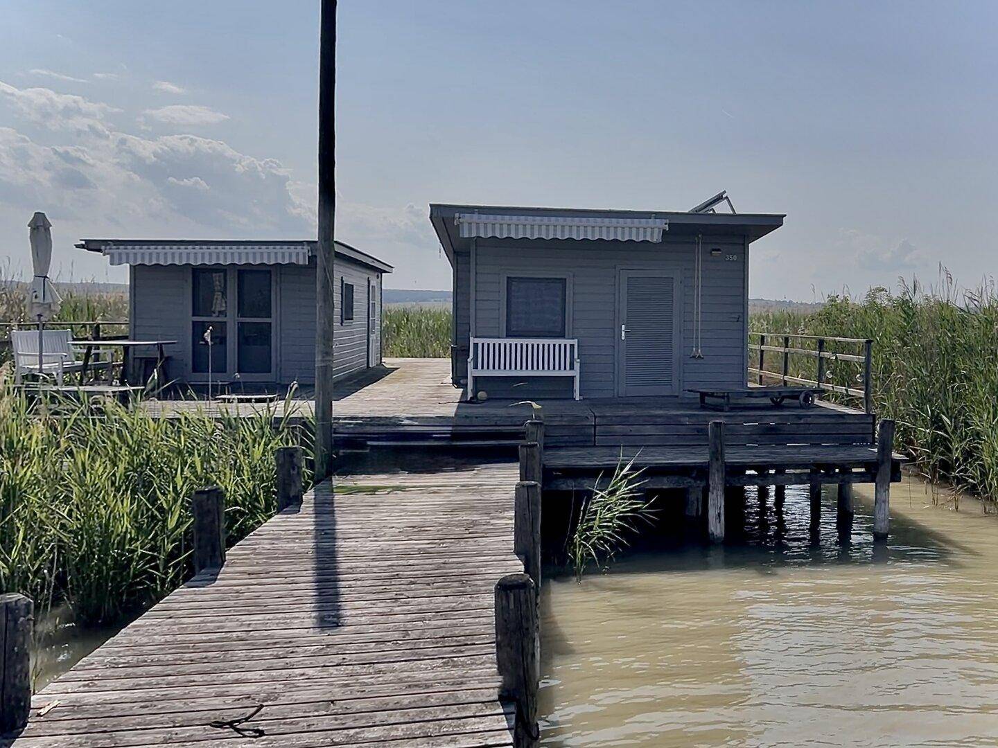 Die beiden kleinen Häuser auf der Holzterrasse am Wasser, umgeben von Schilf und Natur.