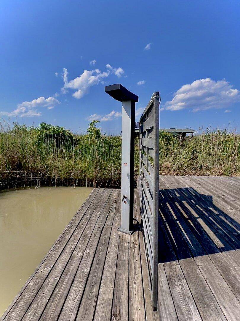 Außendusche auf der Holzterrasse mit Blick auf das Wasser und den blauen Himmel an einem sonnigen Tag.