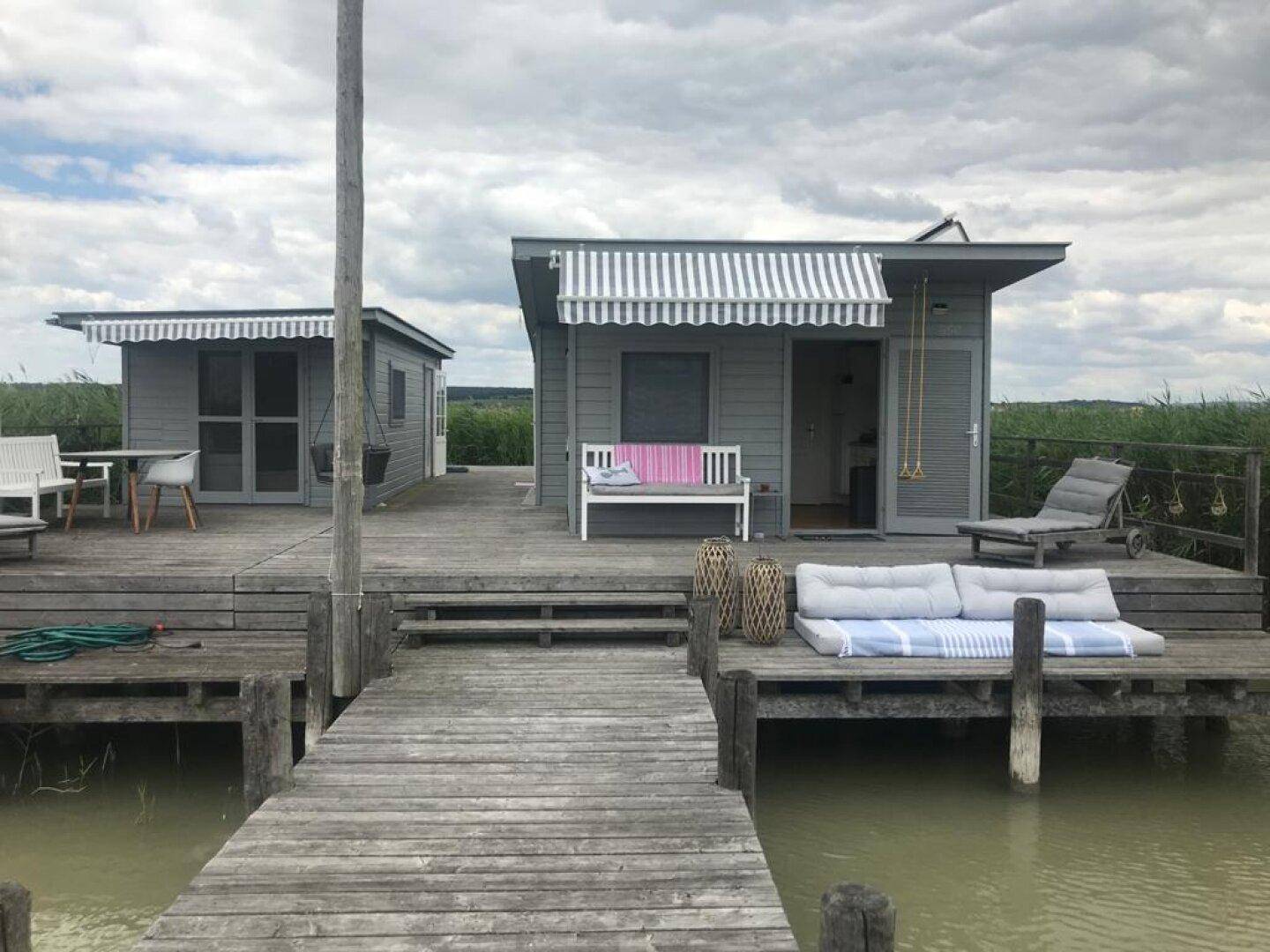 Zwei kleine Häuser auf einer Holzterrasse am Wasser, umgeben von üppigem Schilf und Sitzgelegenheiten.