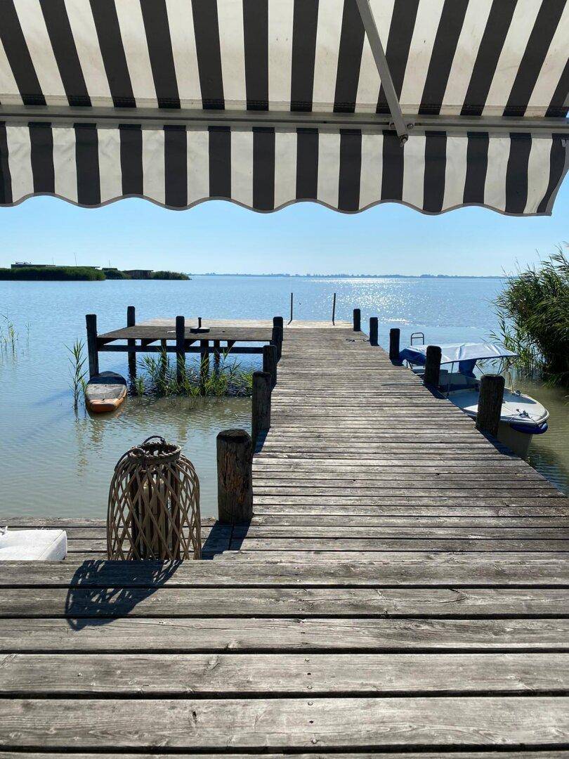 Sonniger Blick von der Holzterrasse auf den ruhigen See unter einer gestreiften Markise.