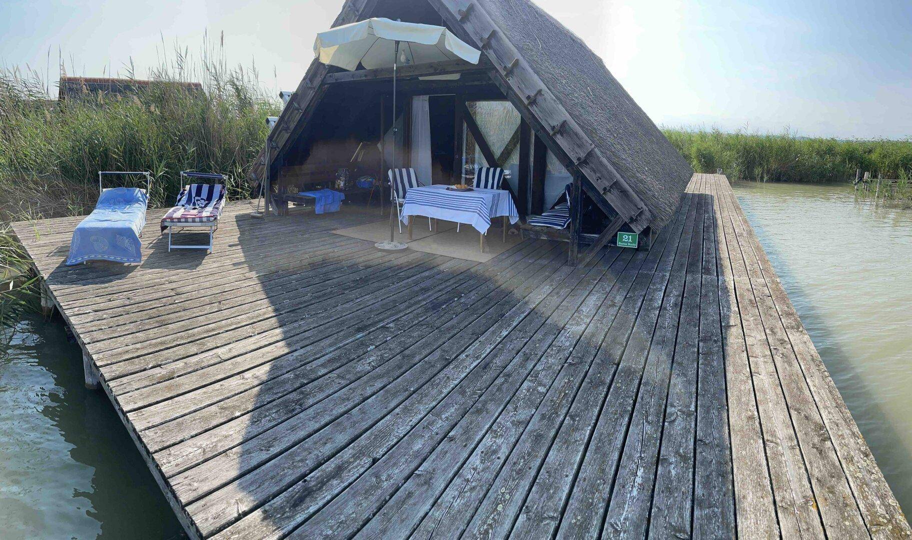 Weitläufige Holzterrasse mit Liegestühlen und Essbereich vor dem Reetdachhaus am See.