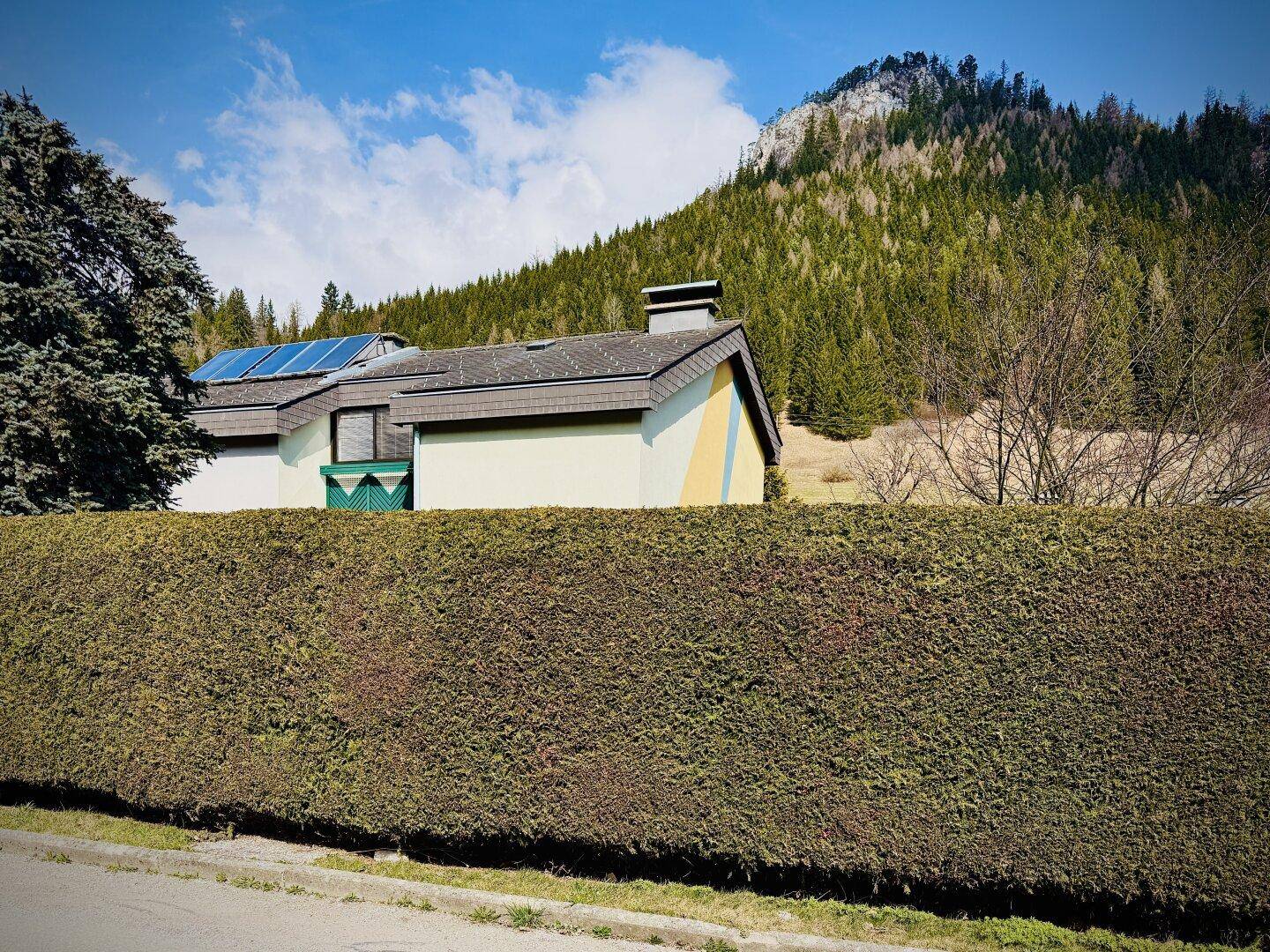 Hausfassade mit Photovoltaikanlage auf dem Dach und umgebender Hecke.