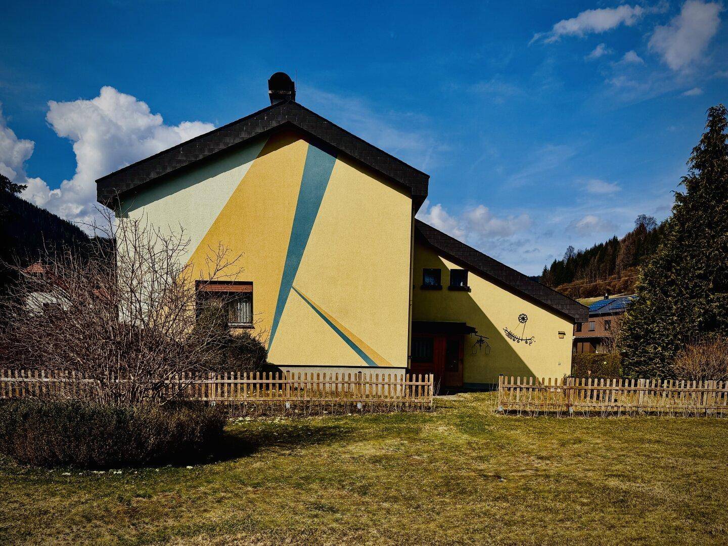 Einfamilienhaus mit gelber Fassade, Holzzaun und weitem Blick auf die Berge.