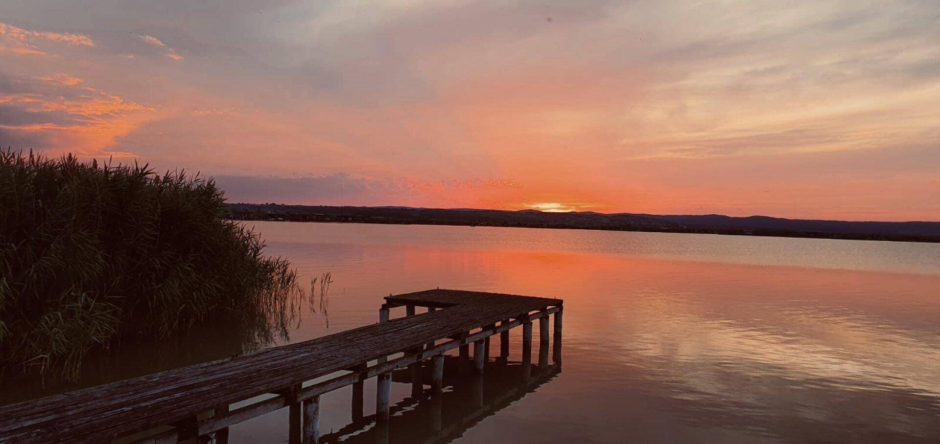 Malerischer Sonnenuntergang über einem See mit einem langen Holzsteg, umgeben von Schilf.