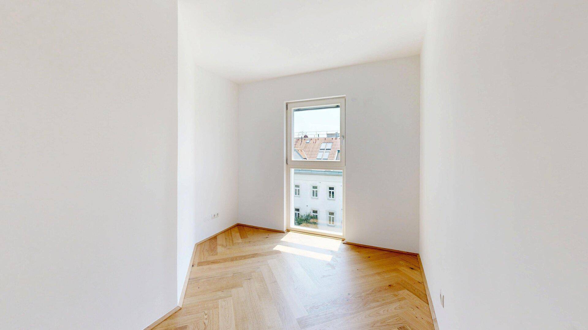 Helles Zimmer mit Fischgrätparkett und Fensterblick auf die umliegenden Gebäude, viel Tageslicht.