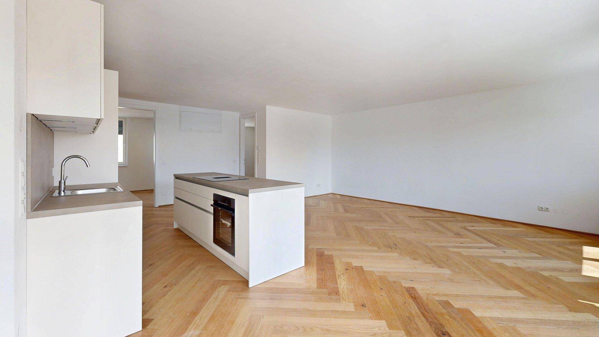 Leerer, offener Wohnbereich mit Kücheninsel und Fischgrätparkett, bereit für individuelle Gestaltung.
