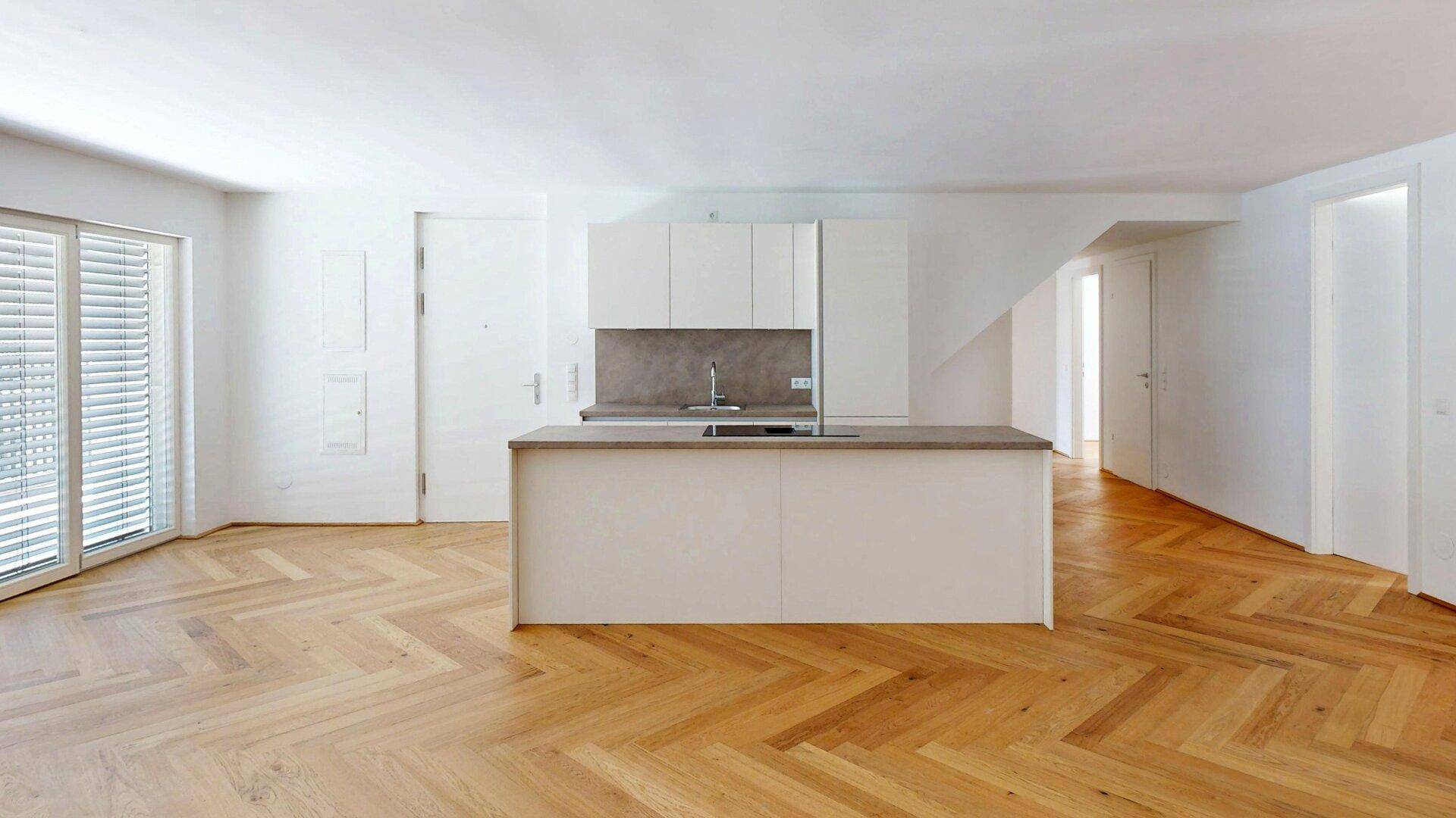 Moderne, leere Küche mit Kücheninsel und Fischgrätparkett, viel Tageslicht durch große Fenster.