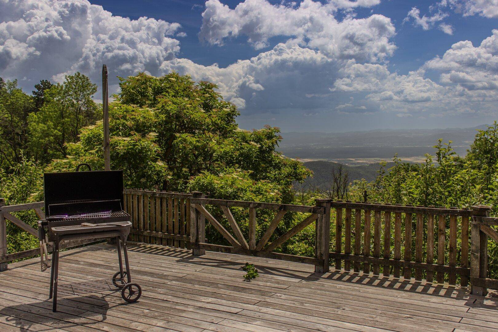 Holzterrasse mit Grill und weitem Blick über die bewaldete Landschaft unter blauem Himmel.