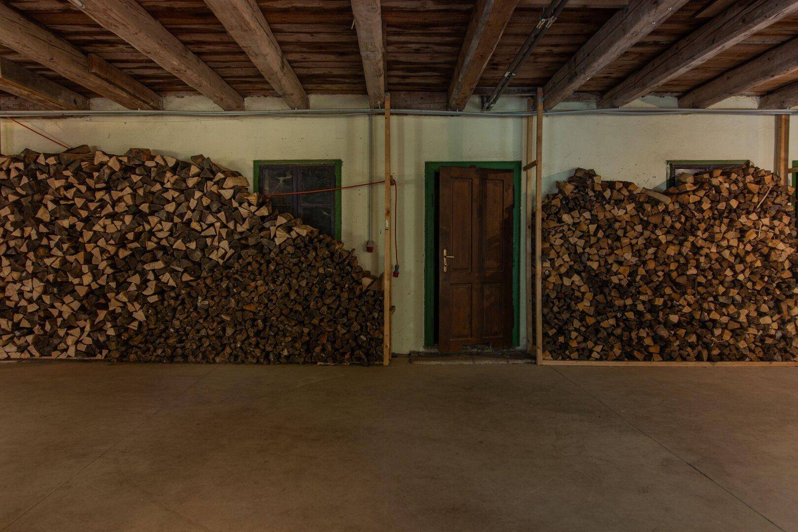 Lagerraum mit großen Holzstapeln, Holztür und kleinen Fenstern.