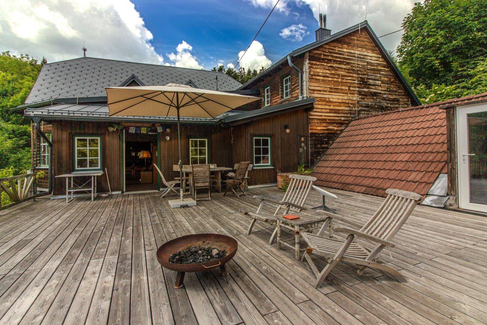 Große Holzterrasse mit Gartenmöbeln, Sonnenschirm und Blick auf das bewaldete Grundstück.