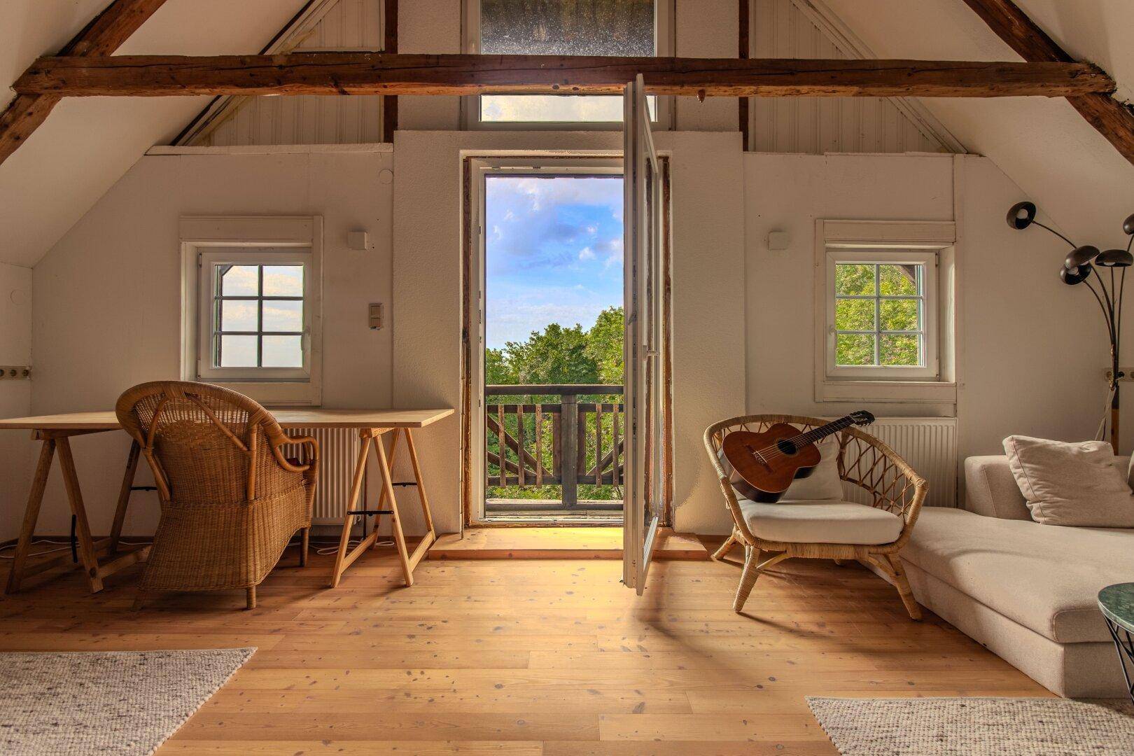 Helles Dachzimmer mit Holzbalken, Holzboden und Zugang zu einem kleinen Balkon.