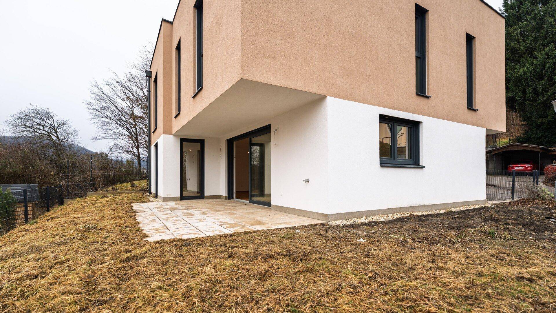Moderne Hausfassade mit einer gepflasterten Terrasse und einem noch ungestalteten Gartenbereich.
