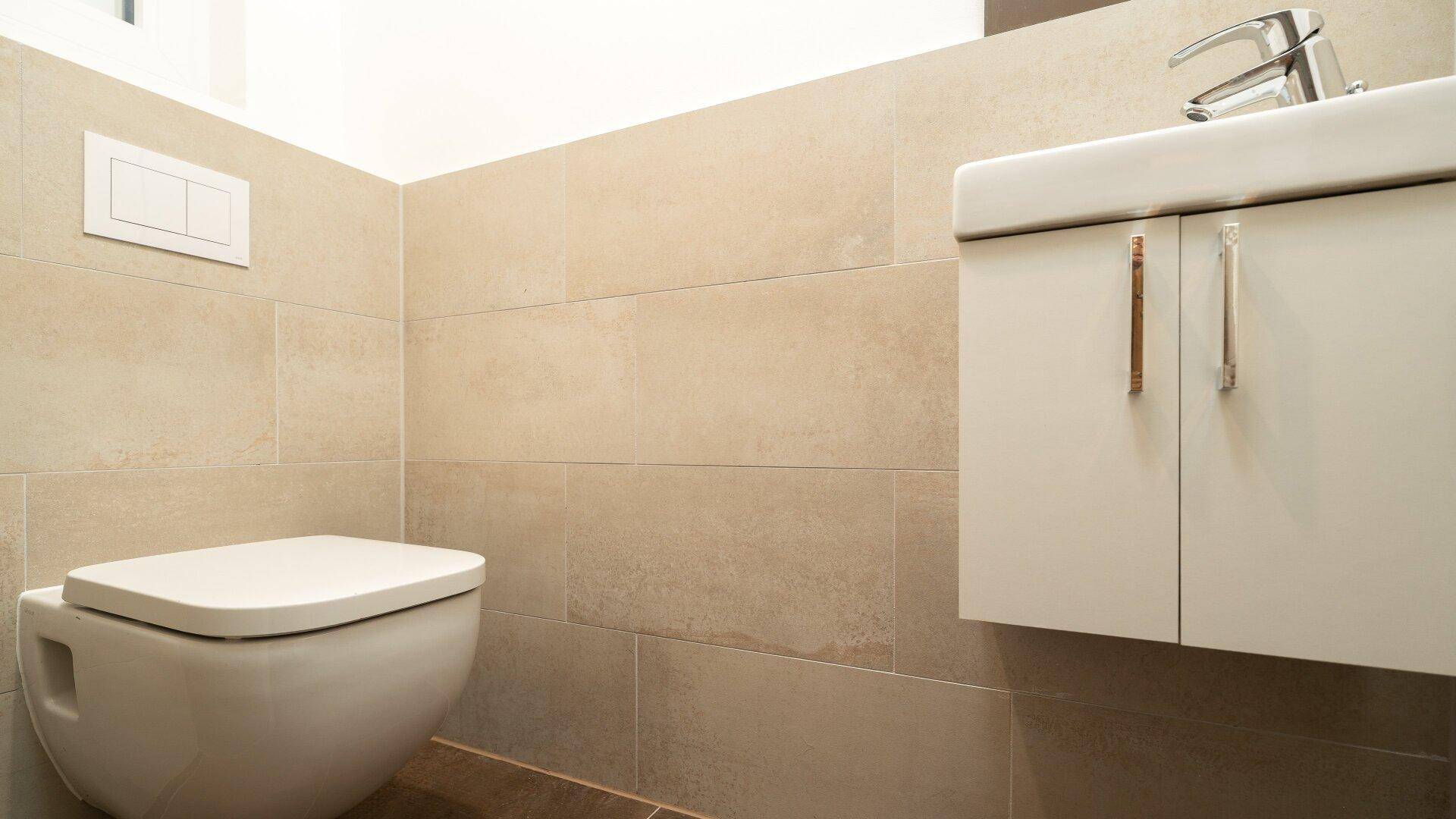 Modernes Gäste-WC mit beigefarbenen Fliesen, wandhängendem WC und einem Waschbecken mit Unterschrank.