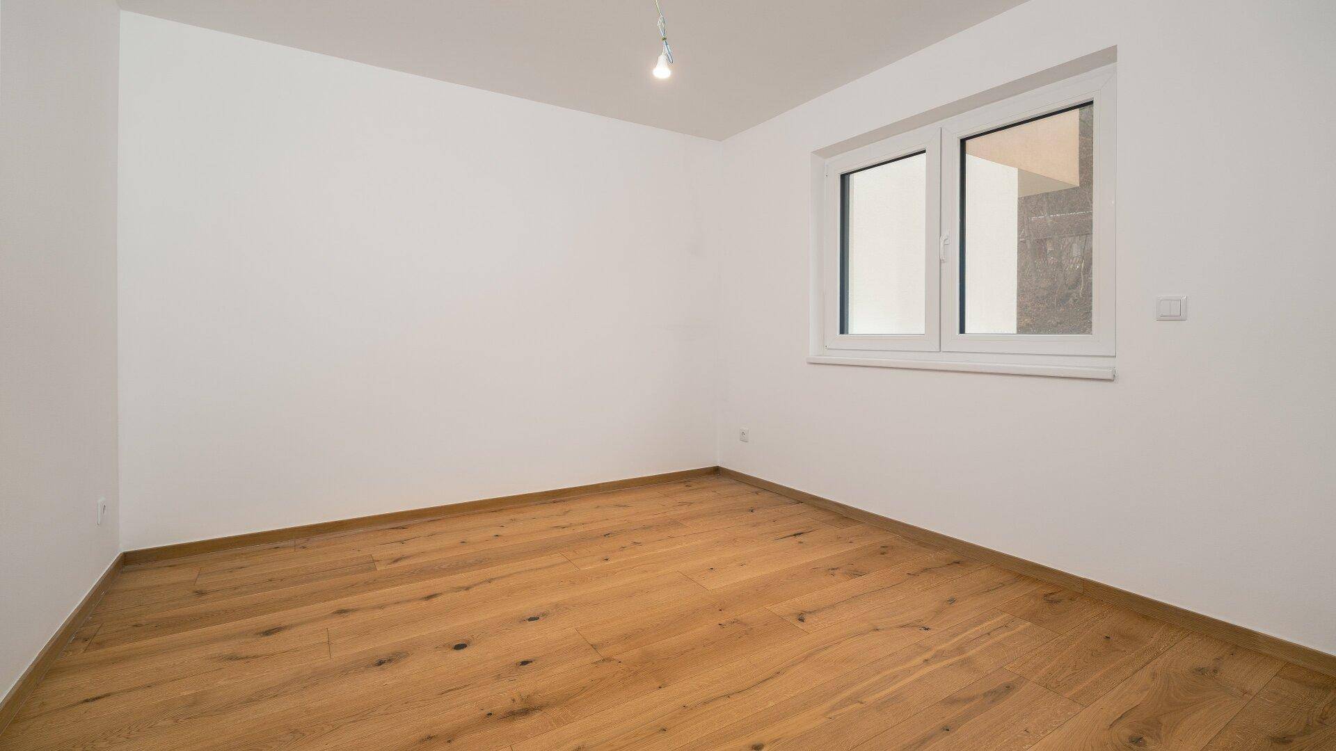 Schlichtes, helles Zimmer mit Holzboden und einem Fenster, bereit zur individuellen Gestaltung.