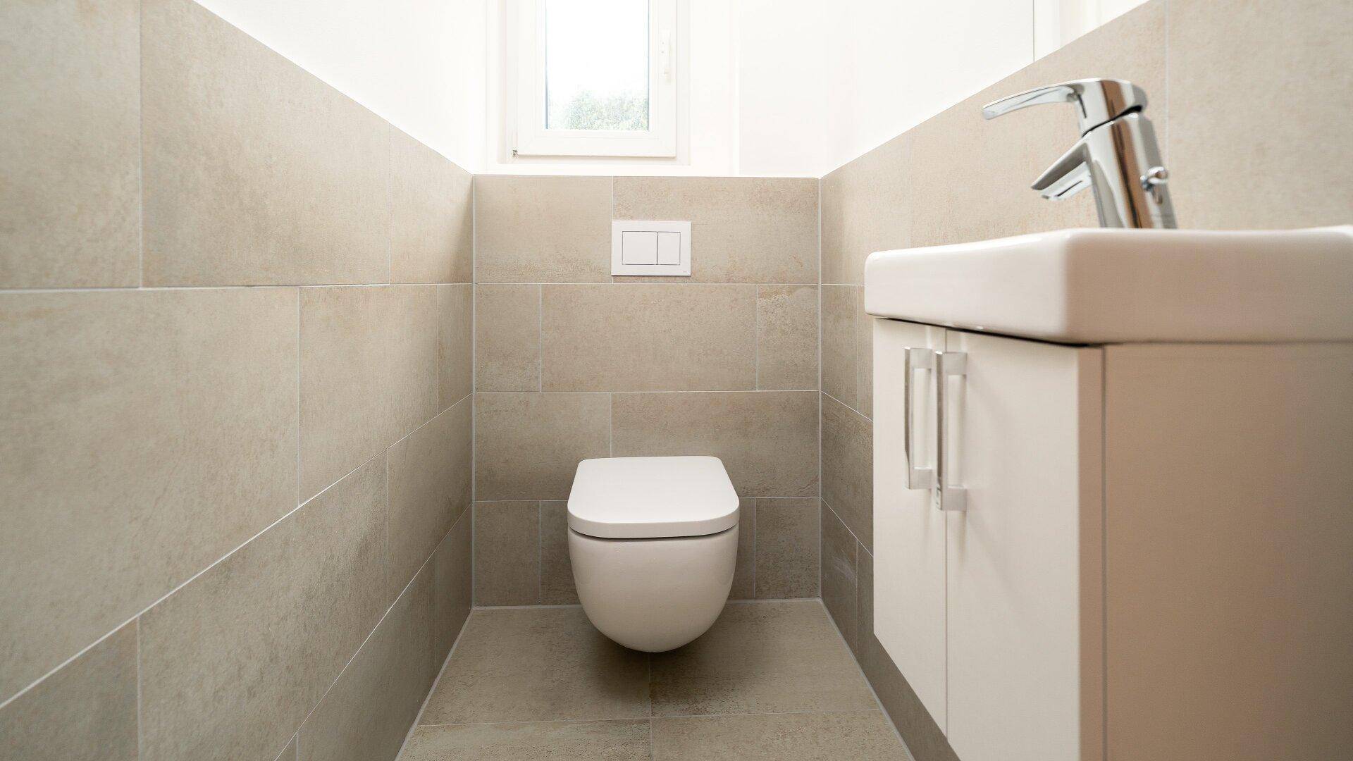 Gäste-WC mit beigefarbenen Fliesen, wandhängendem WC, Waschbecken und einem kleinen Fenster.