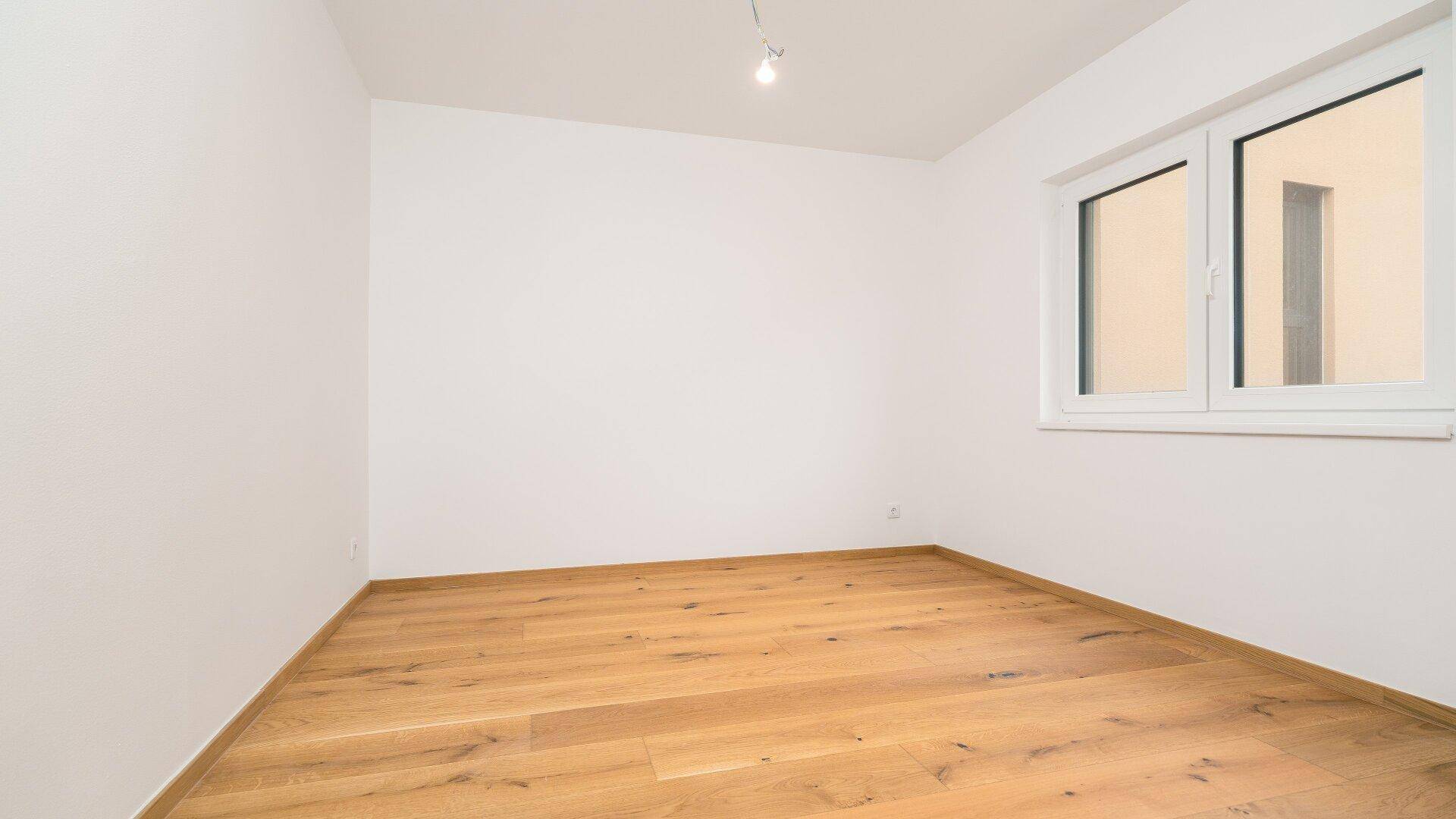 Leeres Zimmer mit Holzboden und einem Fenster, das viel Tageslicht in den Raum bringt.