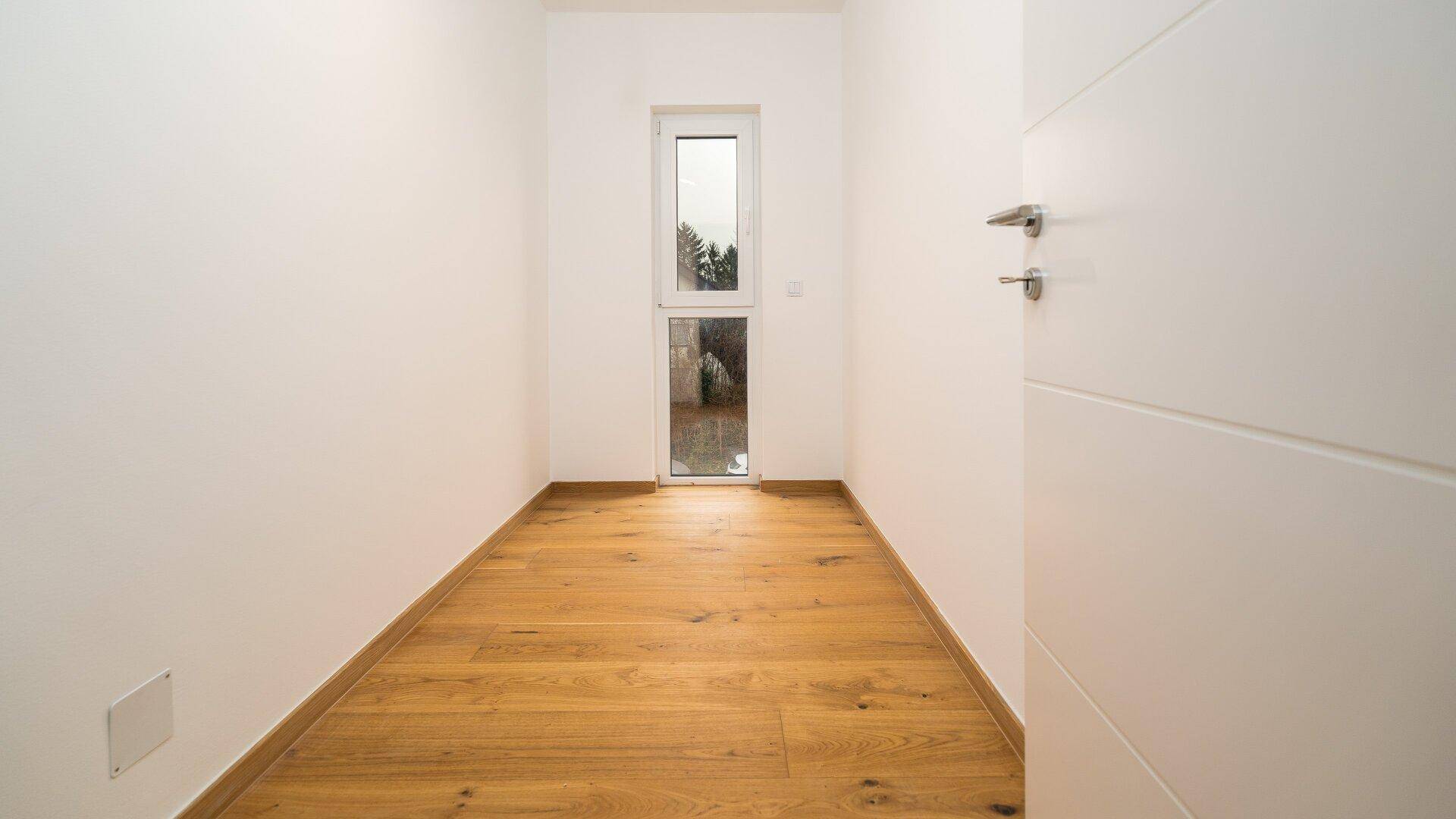 Schmaler Raum mit Holzboden und einem hohen Fenster, ideal als Abstellraum oder Garderobe.