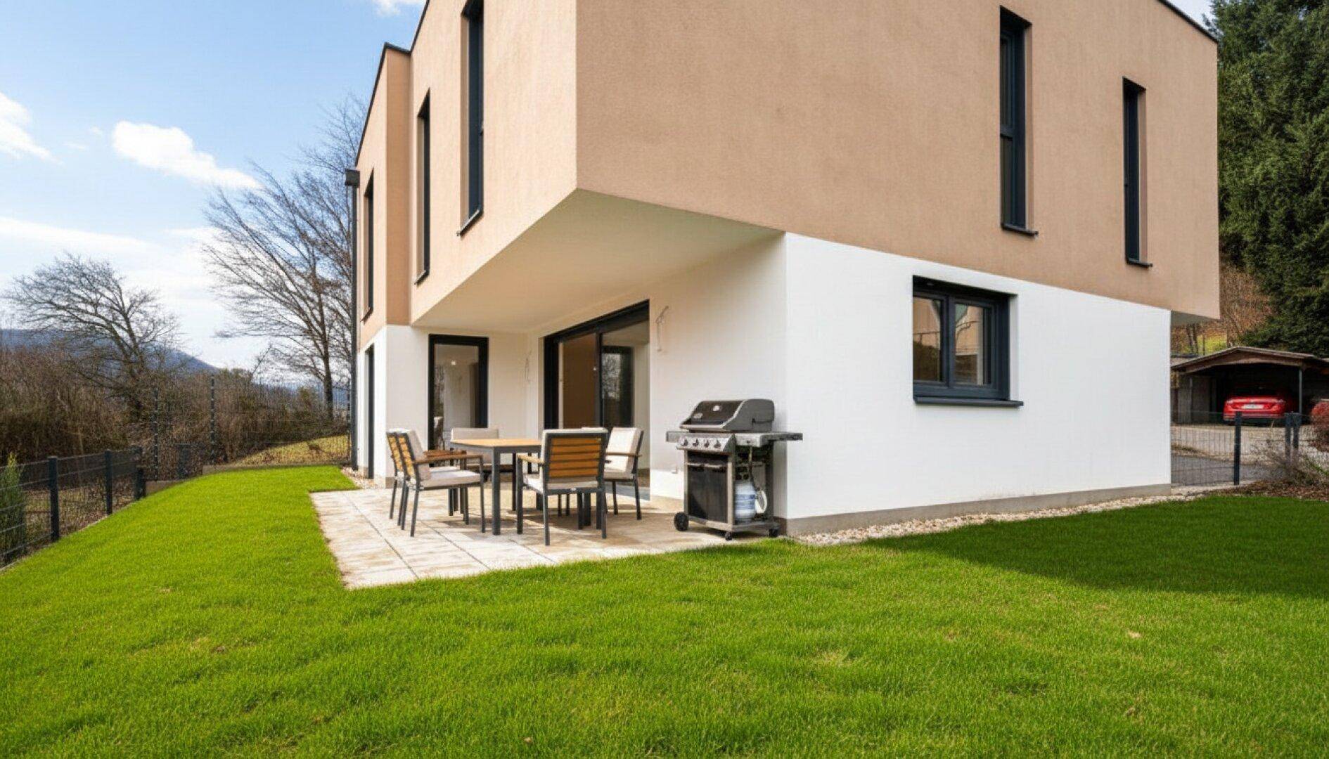 Moderne Hausfassade mit großer Terrasse, Gartenmöbeln und Grillbereich, umgeben von Grün.