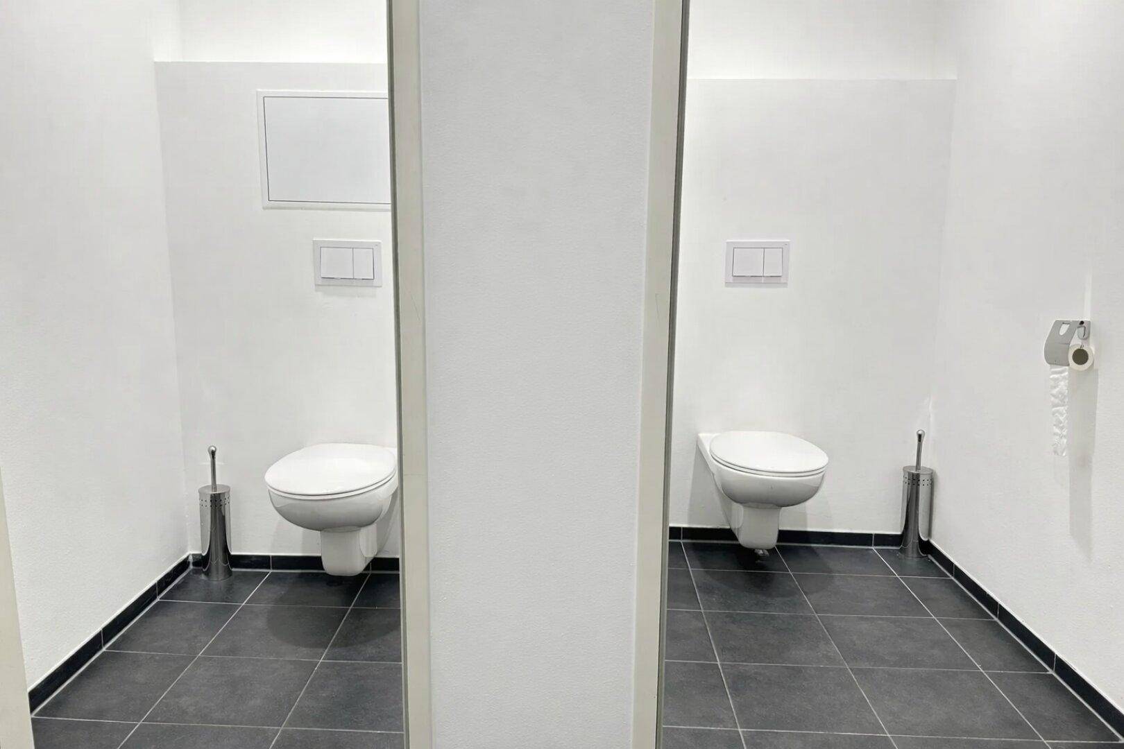 Zwei separate Toilettenräume mit wandhängenden Toiletten und dunklen Bodenfliesen.